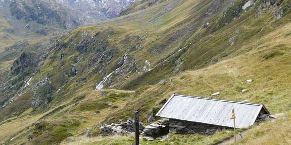 A simple cabin in the picturesque mountain landscape.  
Surrounded by green meadows and tall mountains with snow-capped peaks in the background. - Naturpark Ötztal: Zeitreise um Gurgl -  Wandern auf Ötzis Spuren