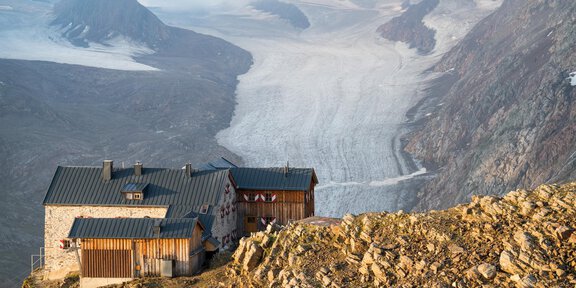 A mountain hut sits on a rocky slope overlooking a glacier. In the background, majestic mountains and a vast glacial landscape can be seen. - 54. Gletscherflohmarsch