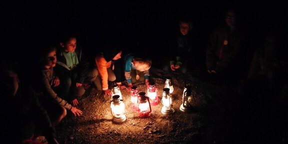 A group of children sits in the dark around several glowing lanterns. The scene conveys a cozy and relaxed atmosphere. - Abenteuer im Laternenlicht