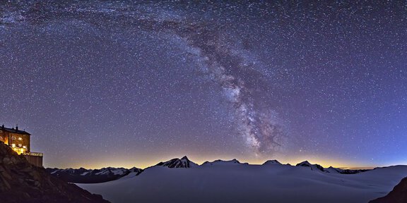 A breathtaking night sky with the Milky Way over snow-covered mountains. In the foreground, a rustic building is visible, illuminated in the dark. - Gemütliches Zusammenkommen unter dem Sternenhimmel
