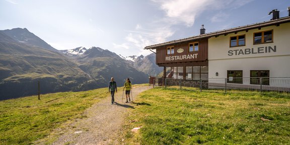 Two people are walking on a path in the mountains. In the background, there is a restaurant with a beautiful mountain landscape visible. -  Gletscher, Geologie & Geschichte – auf Expedition durch das Hochgebirge
