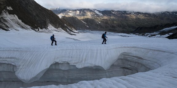 Two mountaineers are walking across a snowy glacier surface. In the background, mountains and a cloudy sky can be seen. - Gletscherspaltentour
