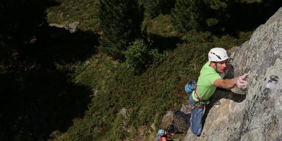 A climber is ascending a rock in a forested environment. In the background, green trees can be seen. - Klettersteig Workshop