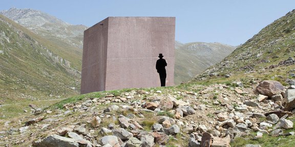 A large gray cube stands in a mountainous landscape. A person near the cube is silhouetted against the impressive backdrop. - Schmugglerwanderung über das Königsjoch (2.825 m)