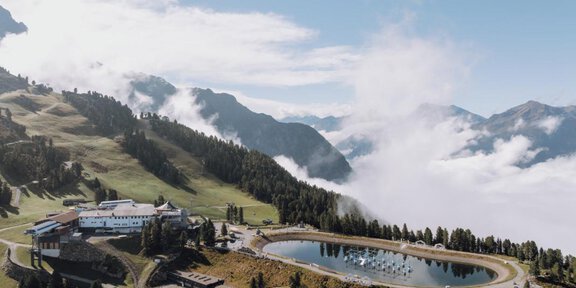 Ein malerischer Bergsee umgeben von grünen Wiesen und majestätischen Bergen. Nebel schwebt über der Landschaft und verleiht der Szene eine mystische Atmosphäre. - Sonnenuntergang am Berg & Fly me into sunset mit Live Musik