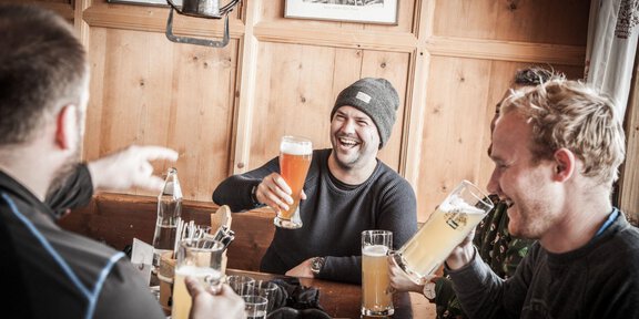 A cheerful group of men is sitting in a cozy bar, lifting beer glasses. They are laughing and enjoying their time together. - Sonnblick - Hochsölden