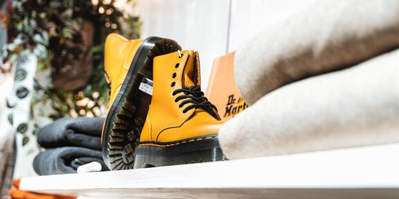 Ein Paar auffällige gelbe Stiefel steht auf einem Regal. Im Hintergrund sind einige Kleidungsteile und Pflanzen zu sehen. - Glanzer Sportboutique | Shop 12 - Sölden