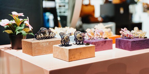 Dekorative Figuren auf einem Tisch, begleitet von kleinen Kunstblumen. Die Objekte sind in verschiedenen Farben und Materialien angeordnet. - Glanzer Sportboutique | Shop 12 - Sölden