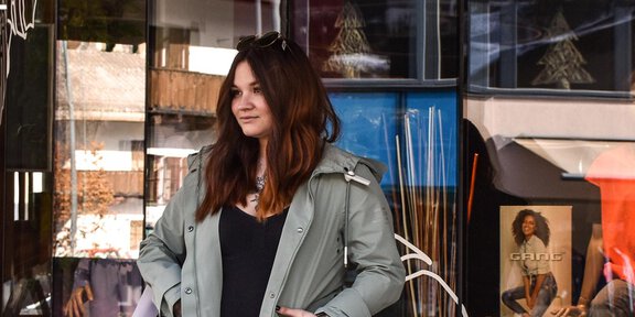 Eine Frau steht vor einem Schaufenster, das mit floralen Mustern dekoriert ist. Sie trägt einen hellgrünen Mantel und sieht entspannt aus. - Glanzer Sportboutique | Shop 12 - Sölden