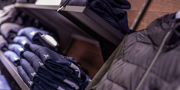 Eine Auswahl an gefalteter Sportkleidung in dunklen Farben auf Regalen. Rechts ist eine schwarze Jacke sichtbar. - THE MOUNTAIN STUDIO - Sölden