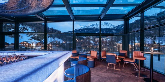 A modern lounge interior with large windows and a view of snow-covered mountains. The cozy furniture and soft light create a relaxed atmosphere. - THE VUE Skybar - Sölden