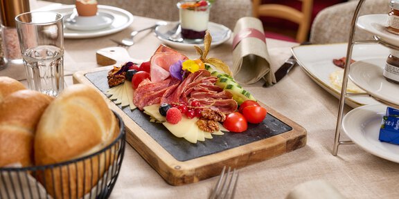 Ein schön angerichteter Teller mit verschiedenen Aufschnittsorten, Tomaten und Trauben. Im Hintergrund sind Brötchen und weitere Speisen auf einem Tisch zu sehen. - Wirtshaus am Giggijoch - Sölden