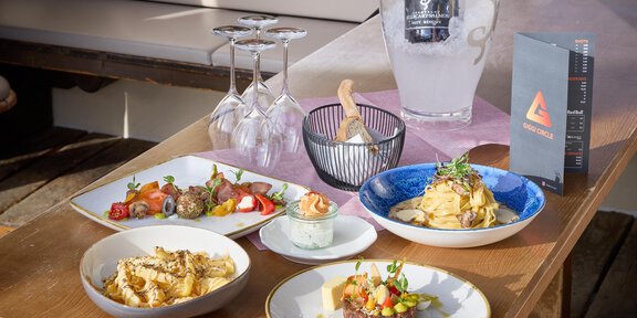 Ein stilvoll gedeckter Tisch mit verschiedenen Gerichten, darunter Pasta, Fleisch und Salat. Im Hintergrund stehen Gläser und eine kühle Flasche Wasser. - Wirtshaus am Giggijoch - Sölden