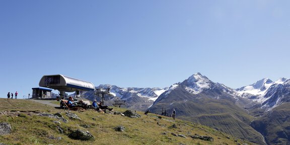 Eine Bergstation mit Menschen inmitten von grünen Wiesen. Im Hintergrund sind schneebedeckte Berge unter einem klaren blauen Himmel zu sehen. - Plattform Wildes Mannle 2.646 m - Vent