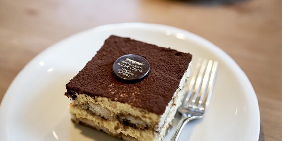 A piece of tiramisu on a white plate with a fork next to it. The cake has a brown cocoa surface and a decorative chocolate plaque. - Schöne Aussicht (Hütte) - Vent