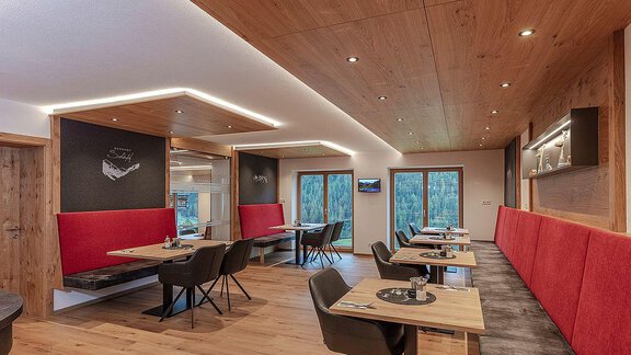 A modern restaurant with wooden decor and red upholstered benches. The tables are elegantly set and offer a view of nature through large windows. - Berghof Schöpf - Gries