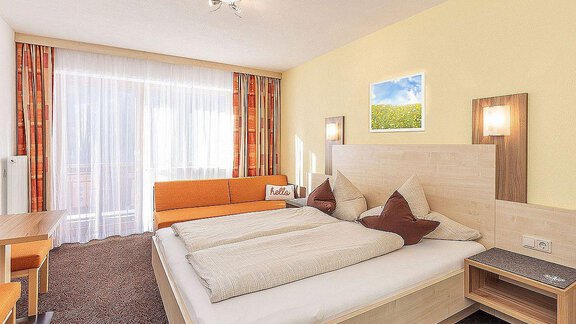 A modern hotel room with a large double bed and bright colors.  
There is a cozy sitting area and plenty of daylight through the windows. - Berghof Schöpf - Gries