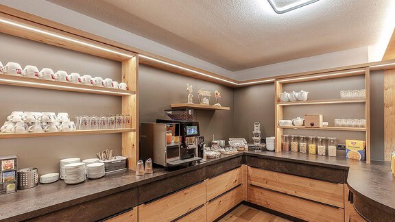 A modern café with wooden shelves and various cups. The counter is neat and offers drinks as well as snacks. - Berghof Schöpf - Gries
