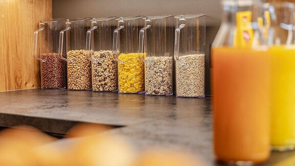 A selection of cereals in transparent containers on a counter. In the foreground are bottles with various juices. - Berghof Schöpf - Gries