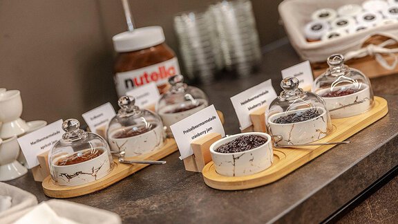 A selection of different spreads in jars on a wooden board. In the background, there is a jar of Nutella. - Berghof Schöpf - Gries