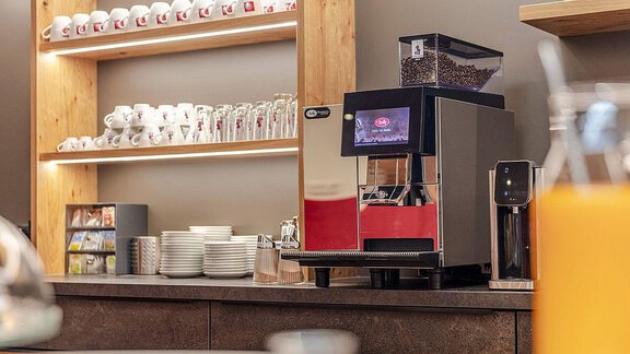 A modern coffee machine stands on a counter next to cups and glasses. In the background, there is a shelf with more coffee cups and snacks. - Berghof Schöpf - Gries