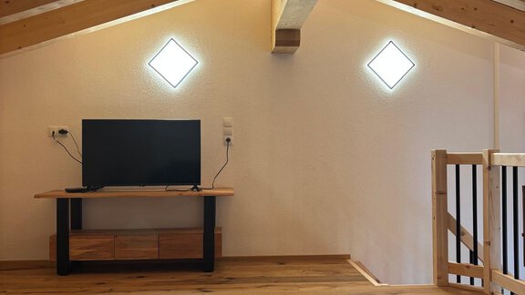 A cozy living room in the attic with a television and a wooden floor. Bright walls and wooden ceilings give the room a warm atmosphere. - KLAMMERHOF - Gries