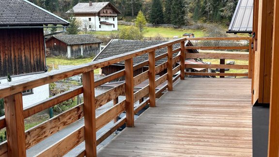 A wooden terrace with railing, surrounded by beautiful mountains and trees. In the background, several houses can be seen. - KLAMMERHOF - Gries