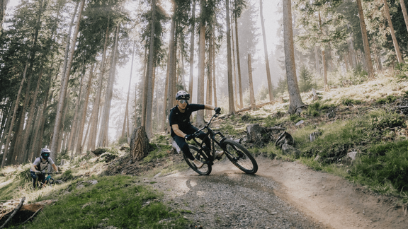 A mountain biker rides quickly on a narrow path through a wooded area. The sun shines through the trees, creating a mystical atmosphere. - Ferienhaus Neurauter - Haiming