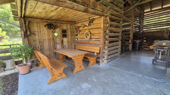 A cozy wooden gazebo with a wooden seating area. The floor is made of concrete and there are some plants in the background. - Landhaus Waldesruh - Haiming
