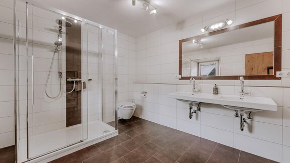 A modern bathroom with a walk-in shower and two sinks. The walls are brightly tiled and the floor is laid with dark tiles. - Landhaus Waldesruh - Haiming