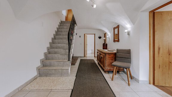 A bright entrance area with a stone staircase. To the right stands a wooden cabinet and a chair. - Landhaus Waldesruh - Haiming
