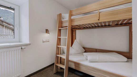 A simple bedroom with a wooden bunk bed. The room has a window and a carpeted floor. - Landhaus Waldesruh - Haiming