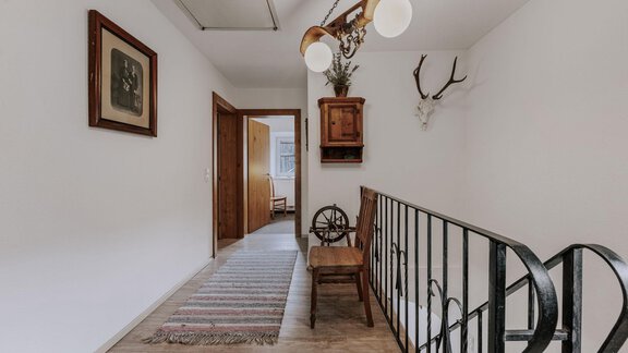 A bright hallway with a wooden railing and a decorative chandelier. On the wall hangs a picture and a deer antler, and in the background, doors to additional rooms can be seen. - Landhaus Waldesruh - Haiming
