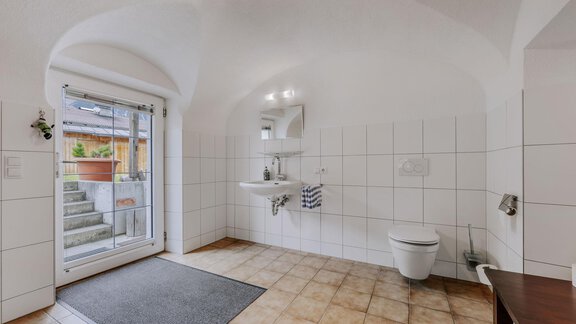 A bright and spacious bathroom with white tiles. There is a sink, a toilet, and access to a terrace through a glass door. - Landhaus Waldesruh - Haiming