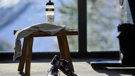 A sports bottle is on a small stool next to a towel and a pair of shoes. In the background, snow-covered mountains and large window light can be seen. - Alpenhotel Laurin - Hochgurgl