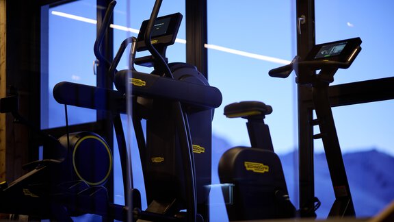 In the gym, various workout machines are placed in front of large windows. The machines are well-lit and offer a view of the mountains. - Alpenhotel Laurin - Hochgurgl