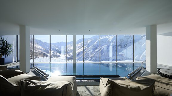 A stylish indoor pool with large windows that offer a view of snow-capped mountains. Cozy seating furniture in the foreground invites you to relax. - Alpenhotel Laurin - Hochgurgl
