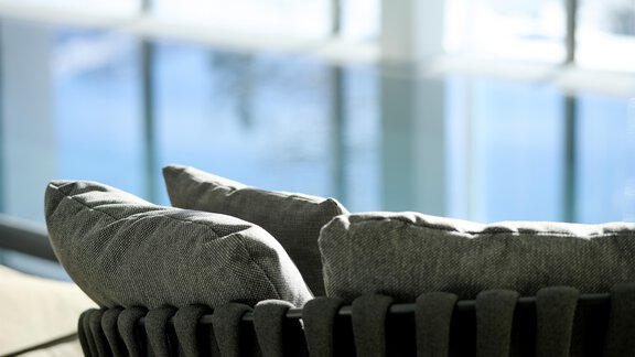 A modern sofa with gray cushions in a bright room. In the background, large windows with a view of water can be seen. - Alpenhotel Laurin - Hochgurgl