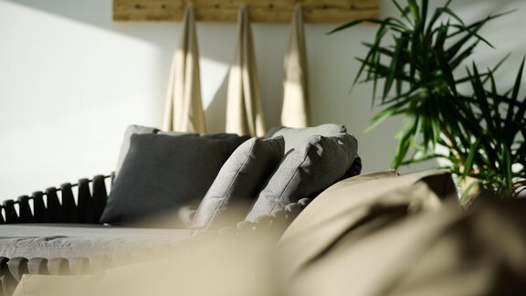A cozy sofa with cushions in a bright room. In the background, some plants and large, bright curtains are visible. - Alpenhotel Laurin - Hochgurgl