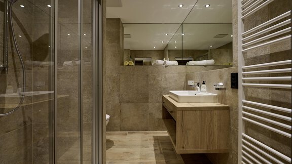 A modern bathroom with a shower and a large mirror. The sink area is made of wood and there are towels on the shelf. - Alpenhotel Laurin - Hochgurgl