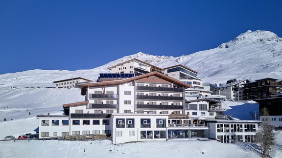 A modern hotel in a snow-covered landscape. In the background, there are mountains and a clear blue sky. - Alpenhotel Laurin - Hochgurgl