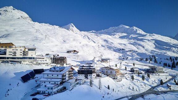 An idyllic winter landscape with snow-covered mountains and a small village. The buildings are surrounded by white splendor and blue sky. - Alpenhotel Laurin - Hochgurgl