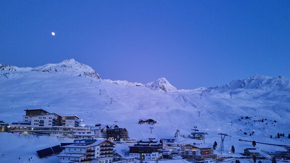 A snowy mountain landscape with a small ski resort. The sky is dark blue and the moon shines over the peaks. - Alpenhotel Laurin - Hochgurgl
