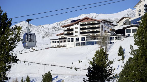 A modern hotel in the mountains, surrounded by snow-capped peaks. A cable car passes by nearby. - Alpenhotel Laurin - Hochgurgl