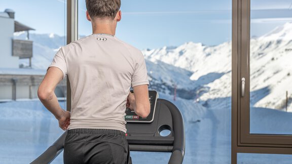 A man is jogging on a treadmill with a view of snow-covered mountains. The room has large windows that let in plenty of daylight. - Hotel Angerer Alm - Hochgurgl