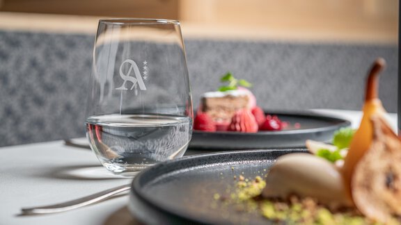 An elegant dish on a black plate, decorated with fruits and a white cream. In the foreground is a glass of water with a modern logo. - Hotel Angerer Alm - Hochgurgl