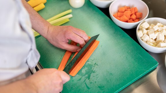 A person is cutting carrots on a green cutting board. In the background, various types of vegetables and bowls with ingredients can be seen. - Hotel Angerer Alm - Hochgurgl