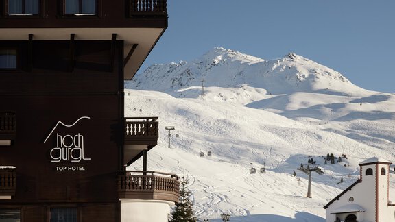 Ein modernes Hotelgebäude mit dem Namen "Hochgugl" in einer verschneiten Berglandschaft. Im Hintergrund sind schneebedeckte Berge und eine Skipiste sichtbar. - TOP Hotel Hochgurgl - Hochgurgl