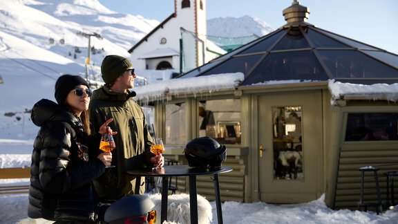 Zwei Personen stehen an einem Tisch im Schnee und genießen Getränke. Im Hintergrund sieht man eine alpine Szene mit einem kleinen Gebäude und Bergen. - TOP Hotel Hochgurgl - Hochgurgl