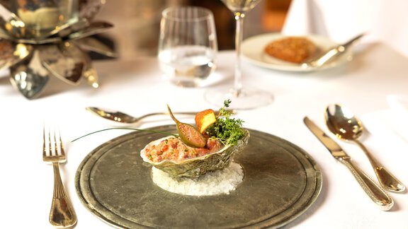 Ein elegantes Gericht auf einem silbernen Teller, verziert mit frischen Kräutern und einer Auswahl an Früchten. Im Hintergrund sind ein Glas Wein und weitere Teller sichtbar. - TOP Hotel Hochgurgl - Hochgurgl